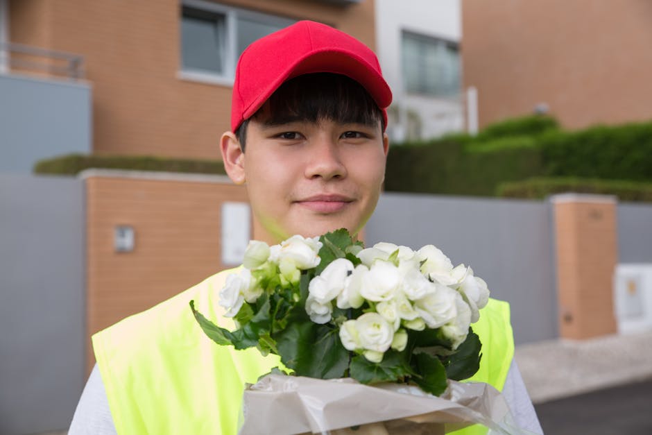 A young delivery person with light skin, dark hair, wearing a red cap and a neon yellow vest, is holding a bouquet of fresh white flowers wrapped in white paper. The bouquet features a cluster of whit