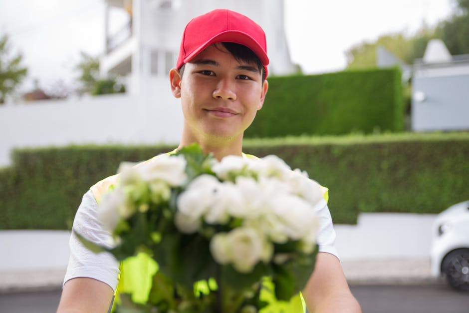 A delivery person wearing a red cap and a high-visibility vest is smiling and holding a bouquet of fresh white flowers with green foliage, which are wrapped in transparent cellophane. The bouquet is l