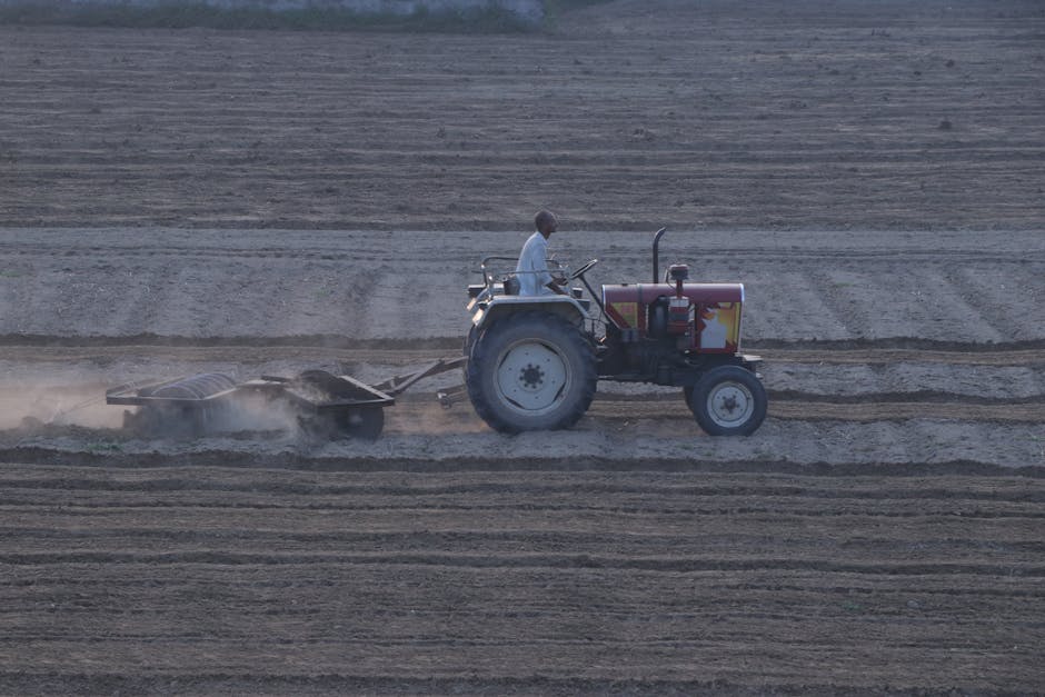 A male farmer wearing a white shirt is seated on a tractor, which is operating on a large, flat, plowed field with visible furrows and dust. The tractor is painted in shades of red and gray and is equ