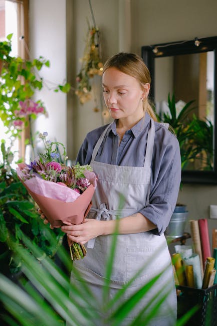 A young woman with light brown hair tied back, wearing a grey button-up shirt and a light grey apron, is holding a bouquet of fresh flowers wrapped in pink paper. The bouquet includes various flowers 