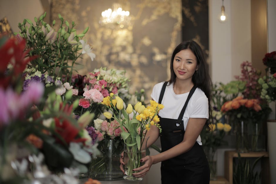 A young woman with long dark hair and a friendly smile, dressed in a white t-shirt and black apron, is arranging a bouquet of fresh yellow and white tulips in a clear glass vase. She is standing in a 