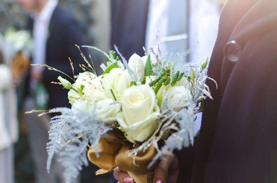 A close-up of a person holding a bouquet of fresh flowers, primarily composed of white roses and white tulips, accented with delicate, wispy foliage and sprigs of lavender. The bouquet is wrapped with