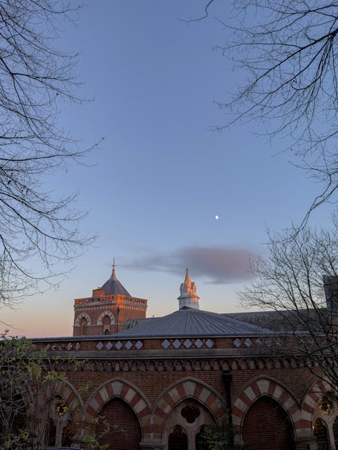 This image shows a view of a historic brick building with ornate red and white arches along the roofline, set against a clear evening sky transitioning from light blue to darker hues, with a visible m