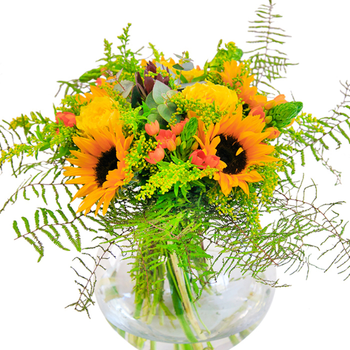 Steeped in the warmth of a Harrow summer, this vivid arrangement captures the spirit of the community in every shining detail. The bouquet's centrepiece is a gathering of radiant sunflowers, their golden faces and textured brown hearts recalling the open, sun-drenched fields just beyond West Harrow. Complementing them are buttery, deeply petalled garden roses, unfolding in elegant whorls and sharing space with bright red Hypericum berries that twinkle like tiny lanterns within the lush green foliage. Layers of feathery ferns and trailing filler greens add softness and natural movement, their textures reminiscent of leafy paths in Harrow-on-the-Hill. Bluish eucalyptus leaves bring an aromatic freshness and silvery accent, enhancing the rustic, garden-fresh feel of the display. Stems are visible through the pristine, round glass vase, elegantly submerged in clear water-a contemporary note that grounds the arrangement with clean lines and clarity. Light streaming across the bouquet highlights the interplay of colour: sunlit yellows, fiery oranges, gleaming greens, and flashes of red, all arranged with the practiced hands of skilled local florists. The scent-delicate yet uplifting-suggests a stroll through Kensington Park in midsummer. Crafted for those milestone occasions-a birthday in Harrow Weald, an anniversary, or simply to brighten the week-this bouquet brings joy, connection, and the unmistakable essence of Harrow's floral artistry into any space.